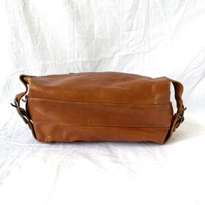 May include: A brown leather shoulder bag with a rectangular shape. The bag has a long strap and metal buckles. The leather has a worn appearance, suggesting it is vintage or has been well-used. The bag is set against a white background.