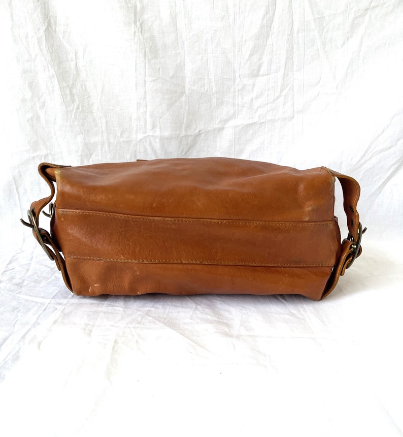 May include: A brown leather shoulder bag with a rectangular shape. The bag has a long strap and metal buckles. The leather has a worn appearance, suggesting it is vintage or has been well-used. The bag is set against a white background.