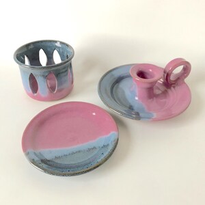 May include: A set of three ceramic pieces in shades of pink and blue. The set includes a small plate, a candle holder with a handle, and a small container with oval cutouts. The pieces have a glazed finish.