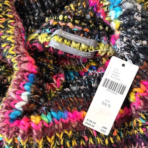 Op de afbeelding: Een veelkleurige gebreide trui met een zwarte achtergrond. De trui heeft een hoge hals en is gemaakt van een dik garen. Het label zegt "By Anthropologie" en "XS/S".