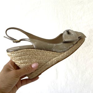 May include: A metallic gold wedge sandal with a bow detail. The sandal features a woven jute wedge heel and a slingback strap with a buckle closure. The shoe is designed for a stylish, comfortable fit.