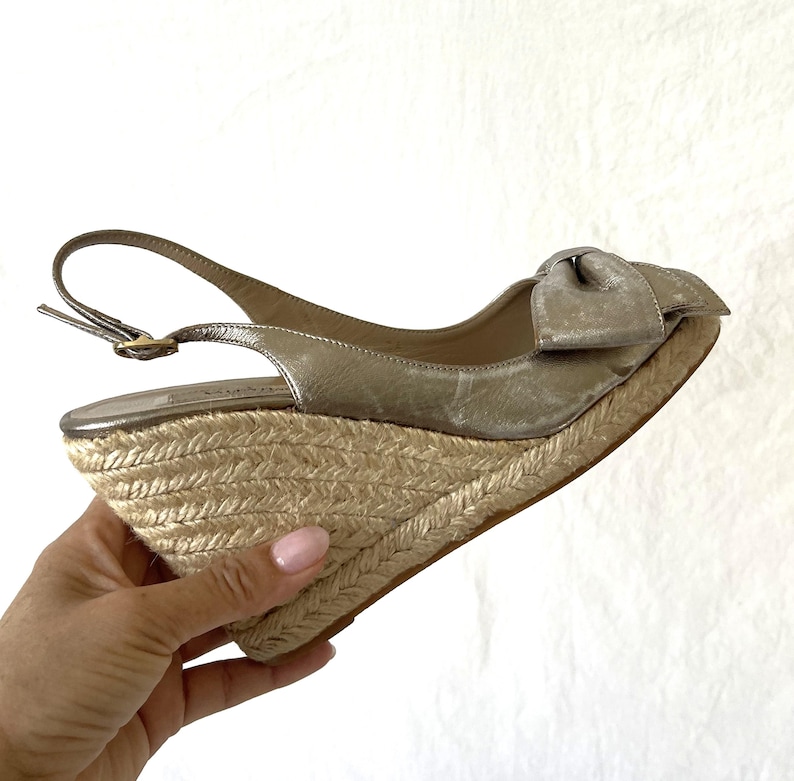May include: A metallic gold wedge sandal with a bow detail. The sandal features a woven jute wedge heel and a slingback strap with a buckle closure. The shoe is designed for a stylish, comfortable fit.