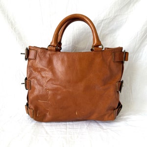 May include: A brown leather tote bag with two top handles and multiple side buckles. The bag has a rectangular shape and a worn appearance, suggesting it is well-used. The background is a white sheet.