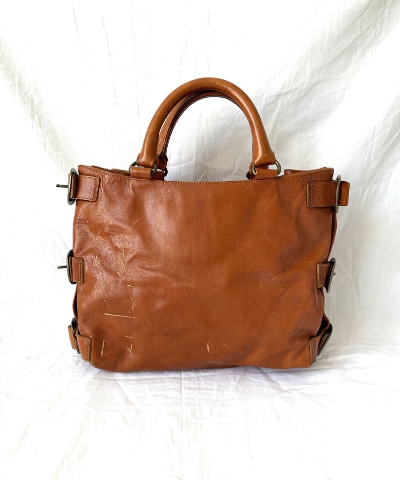 May include: A brown leather tote bag with two top handles and multiple side buckles. The bag has a rectangular shape and a worn appearance, suggesting it is well-used. The background is a white sheet.