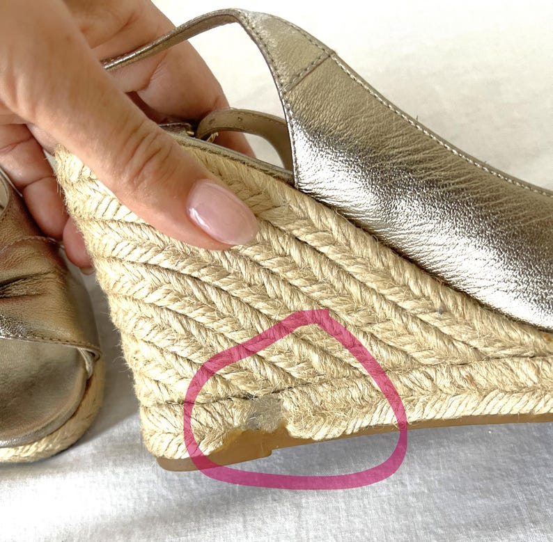 May include: Gold wedge sandals with a woven jute platform and ankle strap. The sandals have a metallic finish and a slingback design. The platform shows some wear and tear. The sandals are a summer footwear item.