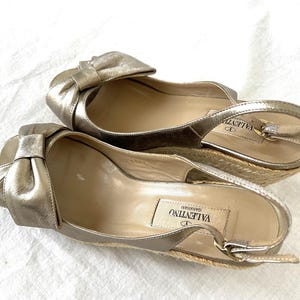 May include: A pair of gold-colored espadrille wedges with a bow detail. The shoes have a slingback strap and a woven jute sole. The Valentino Garavani logo is visible on the insole.