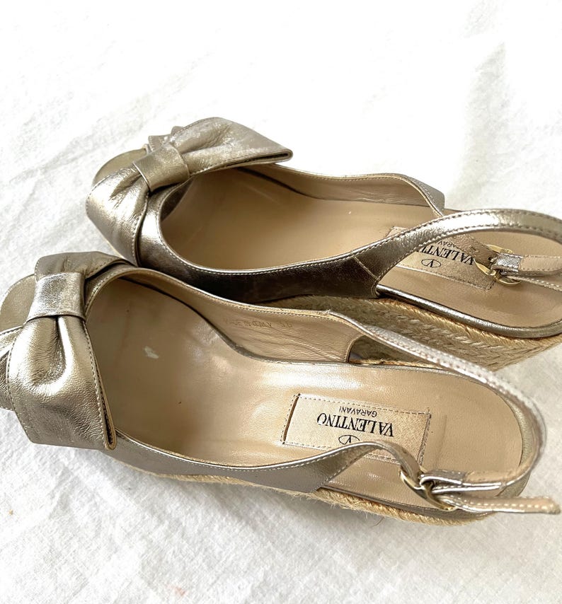 May include: A pair of gold-colored espadrille wedges with a bow detail. The shoes have a slingback strap and a woven jute sole. The Valentino Garavani logo is visible on the insole.