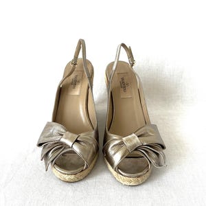 May include: A pair of gold-colored slingback wedge sandals. Each sandal features a large bow detail on the toe, a woven jute sole, and a thin ankle strap. The brand name "Valentino" is visible on the insole.