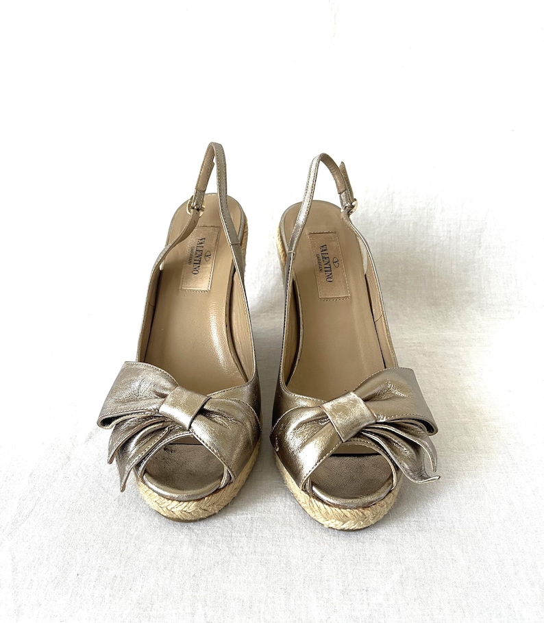 May include: A pair of gold-colored slingback wedge sandals. Each sandal features a large bow detail on the toe, a woven jute sole, and a thin ankle strap. The brand name "Valentino" is visible on the insole.