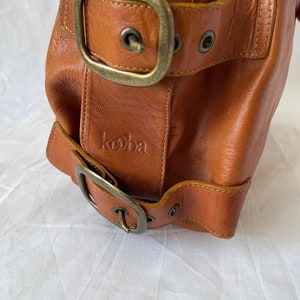 May include: Close-up of a brown leather handbag with two large buckles and a small embossed logo. The bag has a vintage look with visible stitching and antique brass hardware. The leather appears worn and textured.