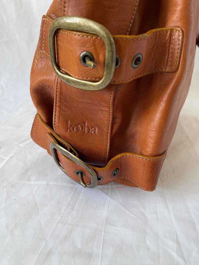 May include: Close-up of a brown leather handbag with two large buckles and a small embossed logo. The bag has a vintage look with visible stitching and antique brass hardware. The leather appears worn and textured.