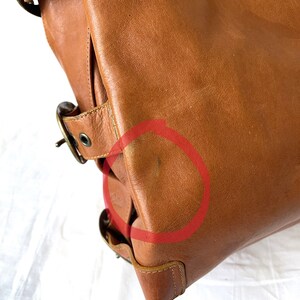May include: A close-up of a brown leather bag with brass-colored buckles and hardware. The bag has a structured design with visible stitching and a slight sheen. The leather shows some wear and tear.