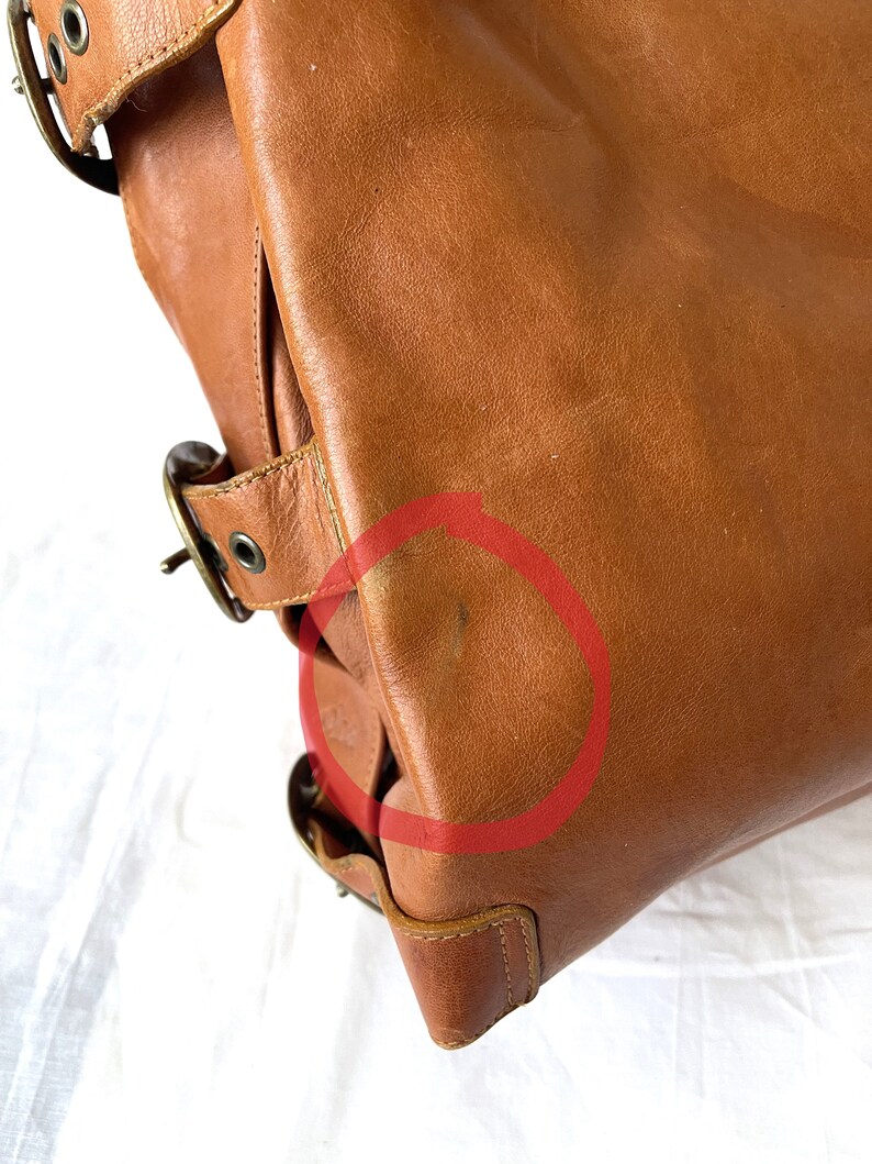 May include: A close-up of a brown leather bag with brass-colored buckles and hardware. The bag has a structured design with visible stitching and a slight sheen. The leather shows some wear and tear.
