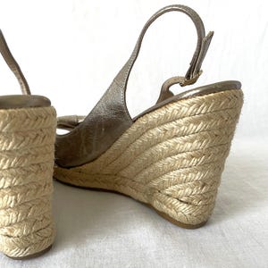 May include: Close-up of a pair of wedge sandals. The sandals feature a metallic gold upper with a slingback strap and a woven jute wedge heel. The wedge heel is approximately 4 inches high.