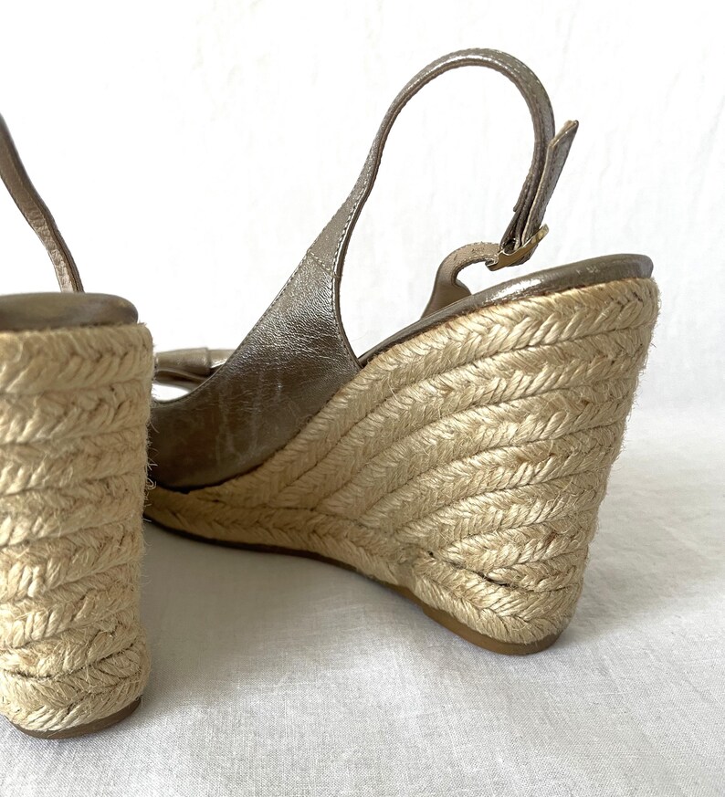 May include: Close-up of a pair of wedge sandals. The sandals feature a metallic gold upper with a slingback strap and a woven jute wedge heel. The wedge heel is approximately 4 inches high.