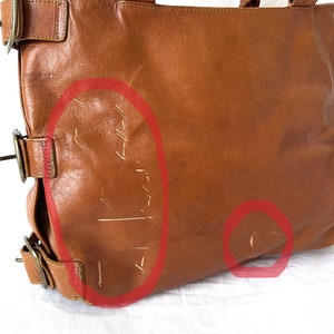 May include: A brown leather tote bag with two handles and metal hardware. The bag shows signs of wear, with scratches and scuffs visible on the surface. The bag has a rectangular shape and a classic design.