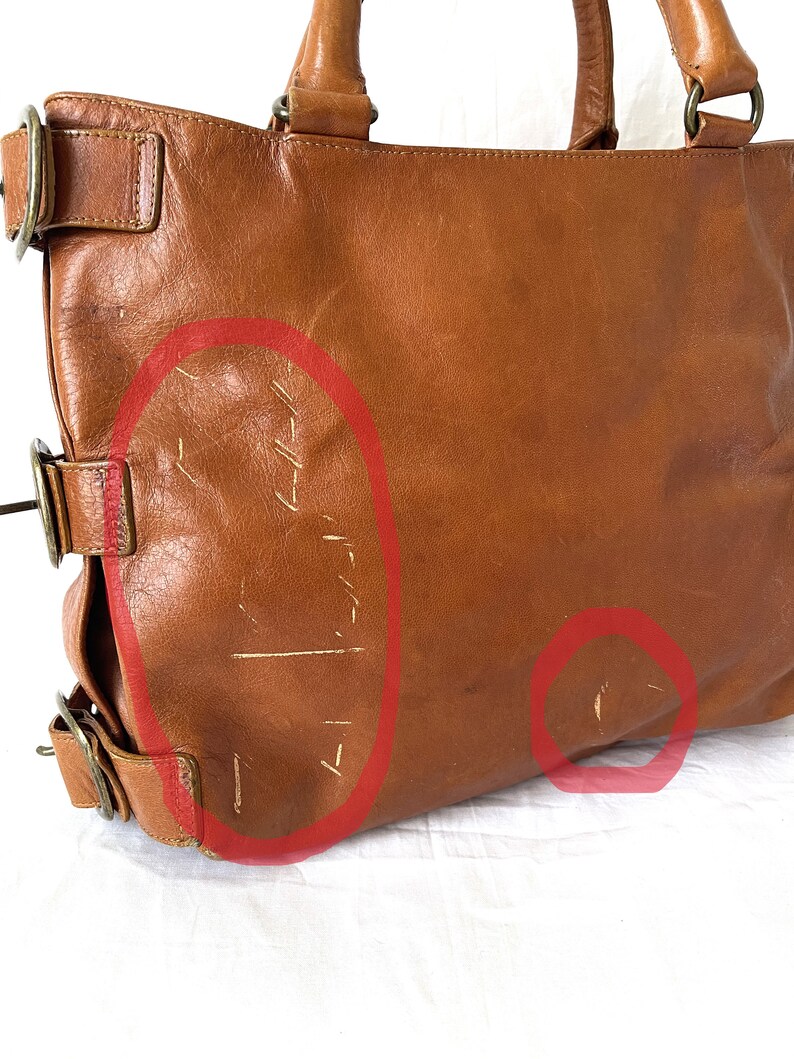 May include: A brown leather tote bag with two handles and metal hardware. The bag shows signs of wear, with scratches and scuffs visible on the surface. The bag has a rectangular shape and a classic design.