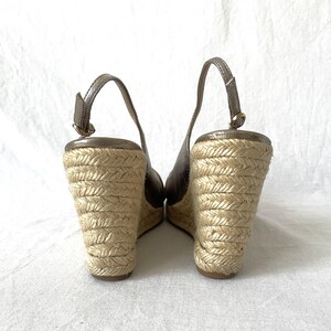 May include: A pair of wedge sandals with a metallic bronze upper and a woven jute sole. The sandals have a slingback strap and a wedge heel. The jute sole is approximately 3 inches high.