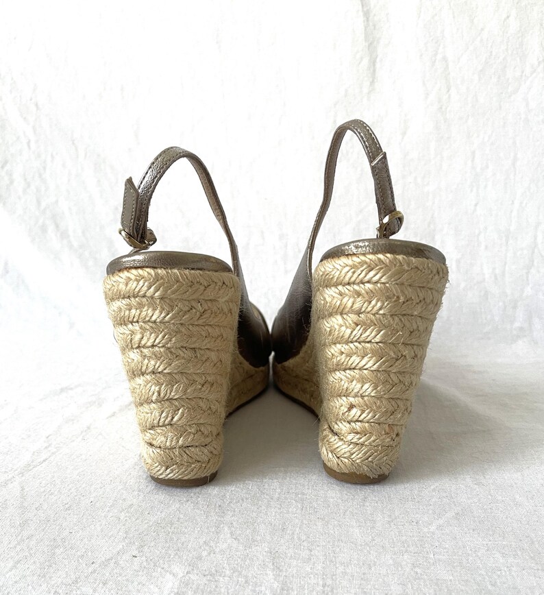 May include: A pair of wedge sandals with a metallic bronze upper and a woven jute sole. The sandals have a slingback strap and a wedge heel. The jute sole is approximately 3 inches high.