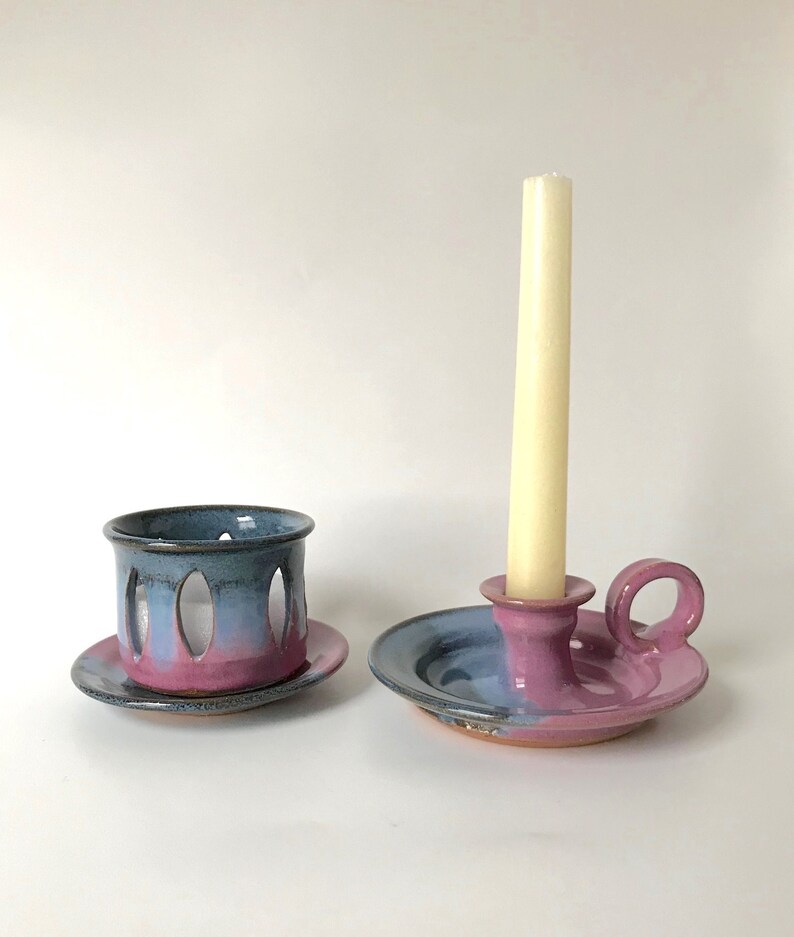 May include: Two handmade ceramic candle holders. One is a small cup with oval cutouts, and the other is a candle holder with a handle. Both are painted with a gradient of blue, pink, and purple.