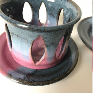May include: A handmade ceramic candle holder with a blue and pink color scheme. The holder has a cylindrical shape with leaf-shaped cutouts and sits on a matching saucer. The glaze has a glossy finish.