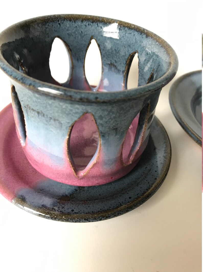 May include: A handmade ceramic candle holder with a blue and pink color scheme. The holder has a cylindrical shape with leaf-shaped cutouts and sits on a matching saucer. The glaze has a glossy finish.