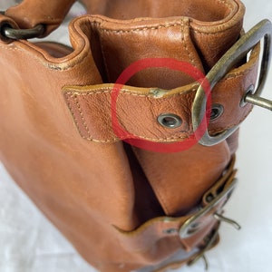 May include: A brown leather handbag with brass-colored hardware. The bag features a structured design with visible stitching and multiple buckles. The leather shows signs of wear, adding to its vintage appeal. The bag has a top handle and a shoulder strap.