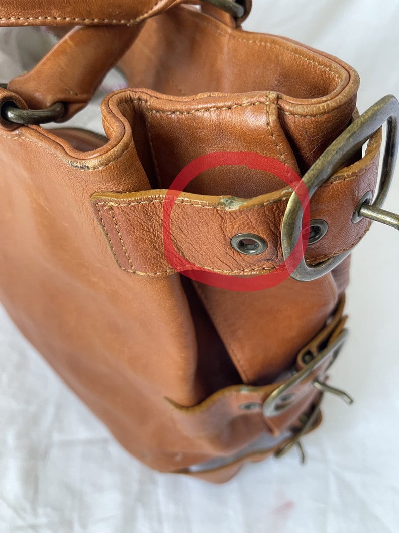 May include: A brown leather handbag with brass-colored hardware. The bag features a structured design with visible stitching and multiple buckles. The leather shows signs of wear, adding to its vintage appeal. The bag has a top handle and a shoulder strap.