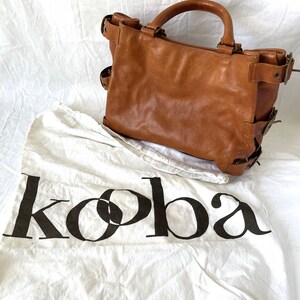 May include: A brown leather tote bag with a top handle and side buckles. The bag sits on a white cloth with the word "koba" printed in black. The bag has a worn, vintage look.