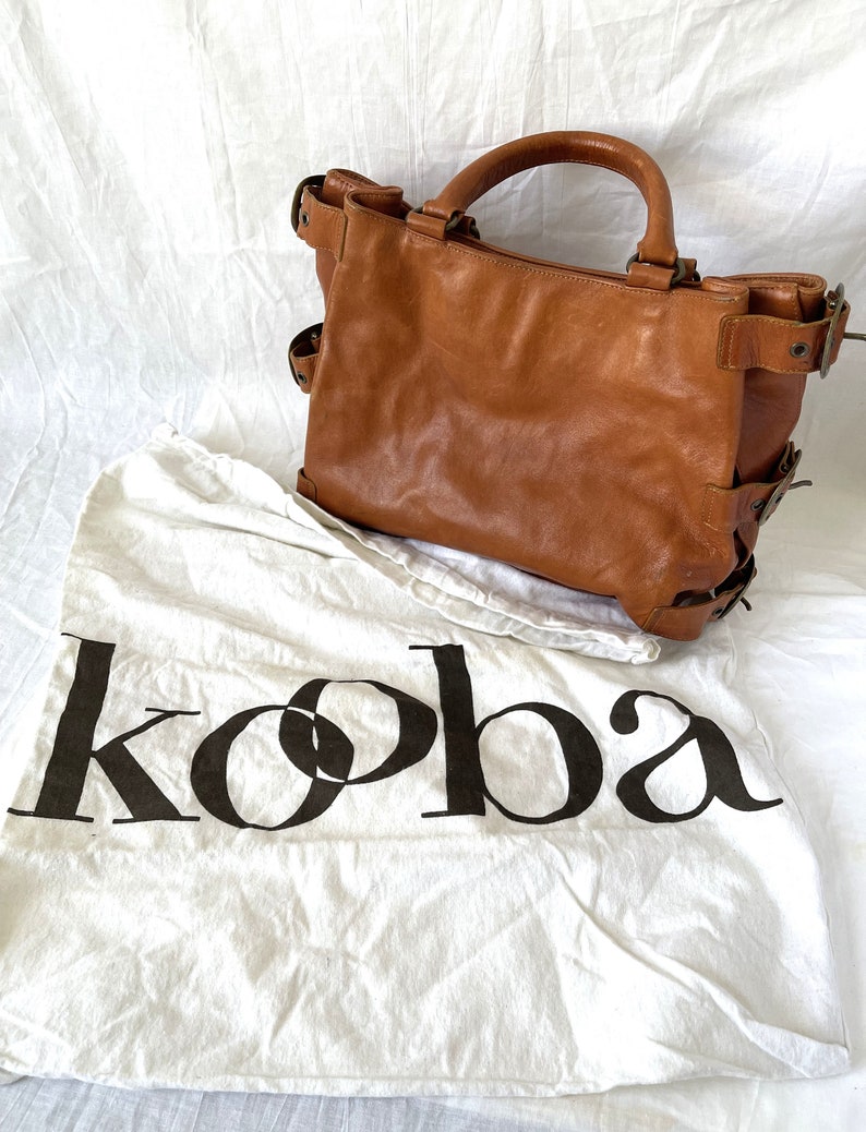 May include: A brown leather tote bag with a top handle and side buckles. The bag sits on a white cloth with the word "koba" printed in black. The bag has a worn, vintage look.