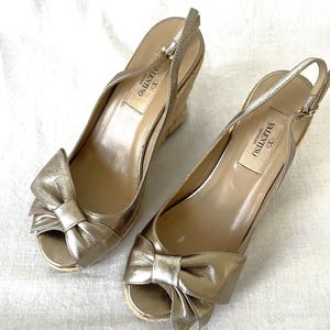 May include: A pair of gold-colored wedge sandals with bow accents. The sandals have a peep-toe design and a woven wedge heel. The brand name "Valentino" is visible on the insole. The sandals are made of a metallic material.