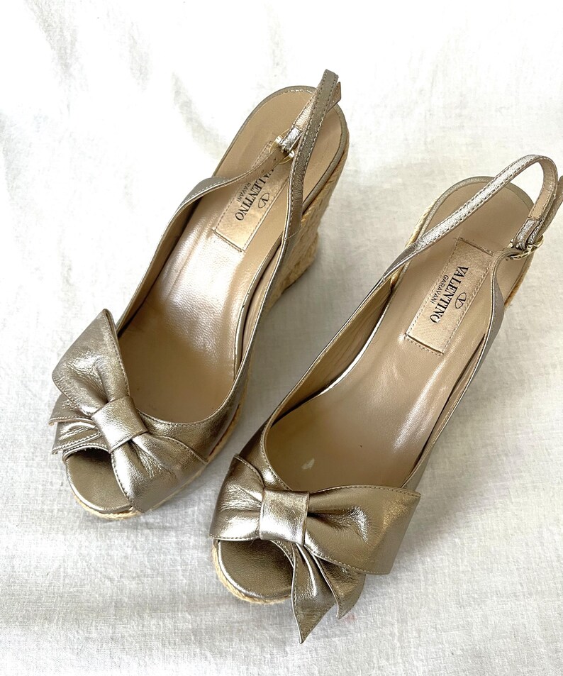 May include: A pair of gold-colored wedge sandals with bow accents. The sandals have a peep-toe design and a woven wedge heel. The brand name "Valentino" is visible on the insole. The sandals are made of a metallic material.