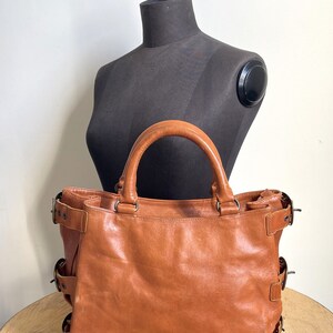 May include: A brown leather tote bag with two handles and multiple buckle details. The bag is displayed in front of a black mannequin. The bag has a worn appearance.