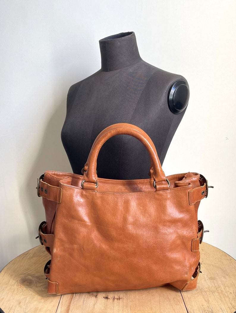 May include: A brown leather tote bag with two handles and multiple buckle details. The bag is displayed in front of a black mannequin. The bag has a worn appearance.