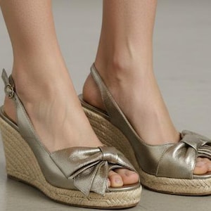 May include: Gold wedge sandals with a bow detail on the toe. The sandals have a woven jute sole and a slingback strap with a buckle closure. The open-toe design is perfect for warm weather.