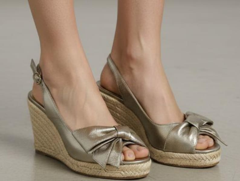 May include: Gold wedge sandals with a bow detail on the toe. The sandals have a woven jute sole and a slingback strap with a buckle closure. The open-toe design is perfect for warm weather.