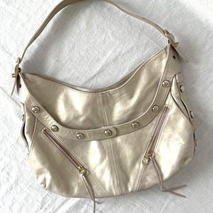 May include: A metallic champagne-colored hobo bag featuring a single shoulder strap, two diagonal zippered pockets with long leather pulls, and a curved decorative panel adorned with six silver-toned metal studs along the front exterior.