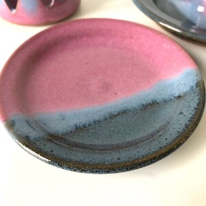 May include: A ceramic plate with a pink and blue glaze. The plate is round with a slightly raised edge. The glaze transitions from pink to blue, creating a two-tone effect. Other ceramic pieces are in the background.
