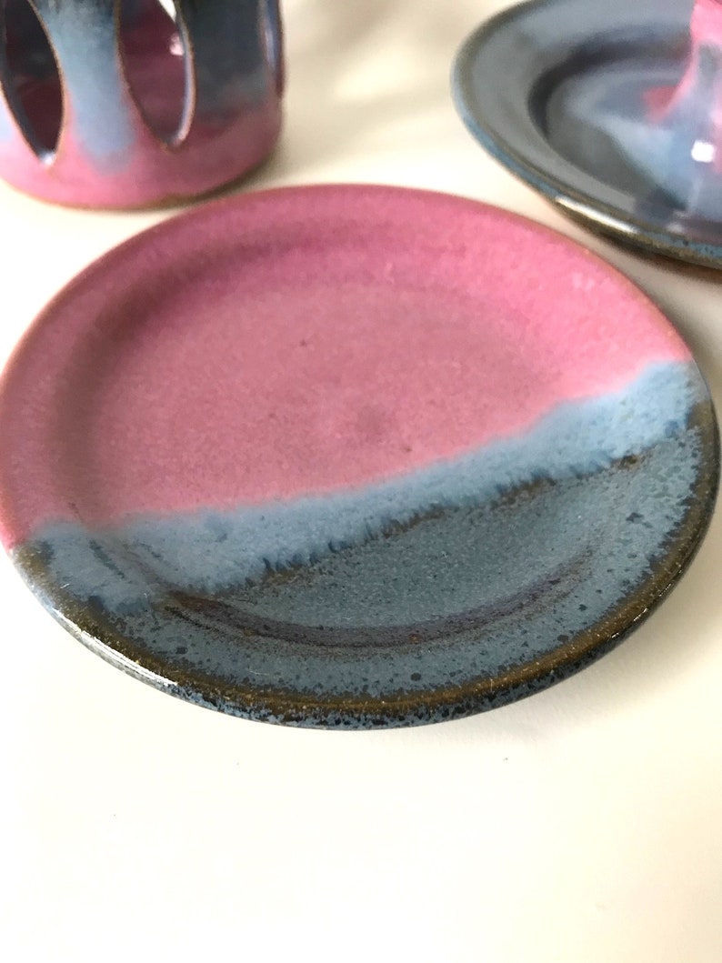 May include: A ceramic plate with a pink and blue glaze. The plate is round with a slightly raised edge. The glaze transitions from pink to blue, creating a two-tone effect. Other ceramic pieces are in the background.