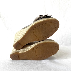 May include: A pair of metallic gold wedge sandals with a bow detail. The soles are brown, and the wedge heels are wrapped in a woven, natural-colored material. The size 36 is visible on the sole.