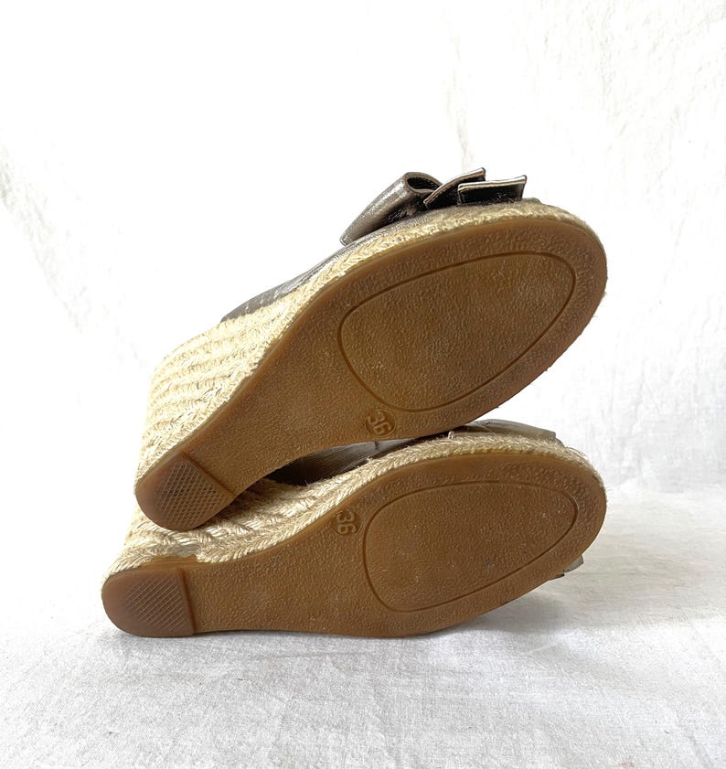 May include: A pair of metallic gold wedge sandals with a bow detail. The soles are brown, and the wedge heels are wrapped in a woven, natural-colored material. The size 36 is visible on the sole.