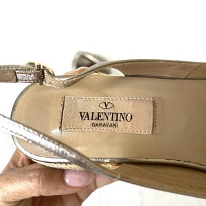 May include: Close-up of a gold-colored Valentino Garavani sandal. The sandal features a woven sole and a thin ankle strap. The Valentino logo is visible on the insole.