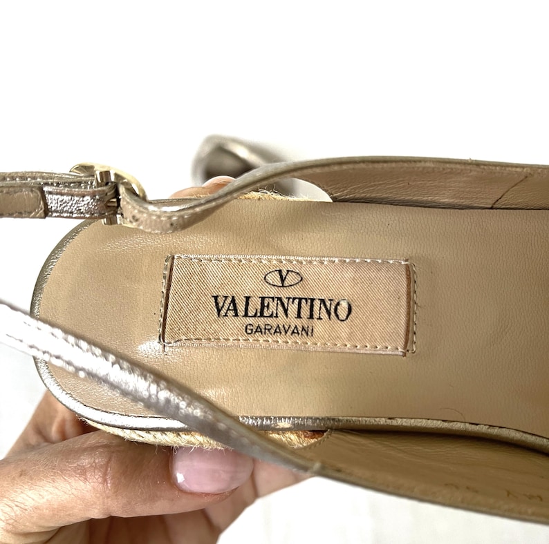 May include: Close-up of a gold-colored Valentino Garavani sandal. The sandal features a woven sole and a thin ankle strap. The Valentino logo is visible on the insole.