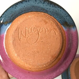 May include: A handmade ceramic plate with a unique color scheme. The plate features a vibrant pink rim that transitions to a deep blue edge. The bottom of the plate is a warm, earthy brown with the word "Wagon" inscribed.