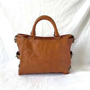 May include: A brown leather tote bag with two handles and metal hardware. The bag has a rectangular shape with side buckles and a spacious interior. The leather has a smooth texture and a warm, natural color.