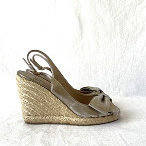May include: Gold wedge sandals with a bow detail. The sandals have a woven jute wedge heel and a slingback strap. The open-toe design and metallic finish create a stylish look. The heel height is approximately 4 inches.