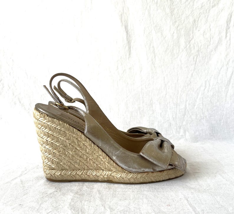 May include: Gold wedge sandals with a bow detail. The sandals have a woven jute wedge heel and a slingback strap. The open-toe design and metallic finish create a stylish look. The heel height is approximately 4 inches.