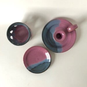 May include: A set of handmade ceramic pottery in shades of pink and blue. The set includes a small plate, a candle holder, and a round container with cut-out holes. The pieces have a glossy finish.