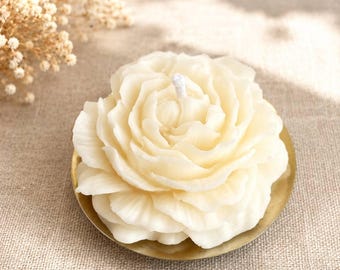 Könnte beinhalten: Eine cremefarbene Kerze in Form einer blühenden Blume mit mehreren geschichteten Blütenblättern und einem einzelnen weißen Docht in der Mitte. Die Kerze steht auf einer kleinen, runden, metallisch goldfarbenen Schale mit glatter, reflektierender Oberfläche.