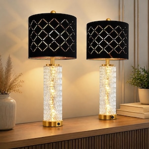 May include: A pair of table lamps featuring cylindrical clear textured glass bases with internal warm lighting and black drum shades. The shades have a geometric diamond cut-out pattern. Each lamp has a gold-toned metal base and a small power switch.